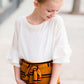 Young girl wearing an ivory, oversized modest top with ruffle sleeve accents. This top is also paired with a mustard, striped below the knee skirt and short boots.