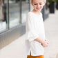 Young girl wearing an ivory, oversized modest top with ruffle sleeve accents. This top is also paired with a mustard, striped below the knee skirt and short boots.