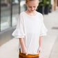 Young girl wearing an ivory, oversized modest top with ruffle sleeve accents. This top is also paired with a mustard, striped below the knee skirt and short boots.