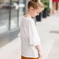 Young girl wearing an ivory, oversized modest top with ruffle sleeve accents. This top is also paired with a mustard, striped below the knee skirt and short boots.