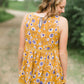 young woman wearing a mustard color tank with blue and white florals. There is embroidered detail just below the chest.