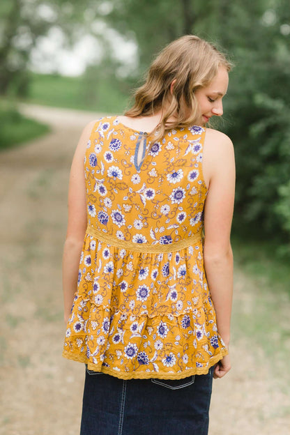 young woman wearing a mustard color tank with blue and white florals. There is embroidered detail just below the chest.