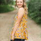 young woman wearing a mustard color tank with blue and white florals. There is embroidered detail just below the chest.