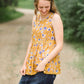young woman wearing a mustard color tank with blue and white florals. There is embroidered detail just below the chest.