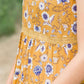 young woman wearing a mustard color tank with blue and white florals. There is embroidered detail just below the chest.