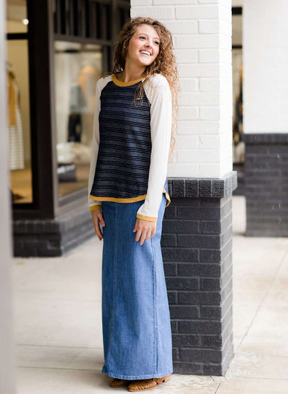 Woman wearing a long denim modest jean skirt. She is wearing a long sleeve, navy raglan top and standing outside of Inherit Clothing Company