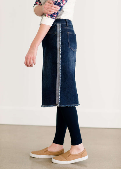 Woman wearing a below the knee jean skirt that has fringe and fray detail on the hem and side seams.