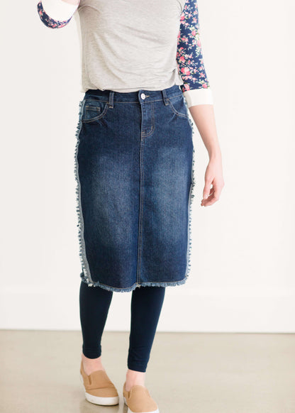 Woman wearing a below the knee jean skirt that has fringe and fray detail on the hem and side seams.