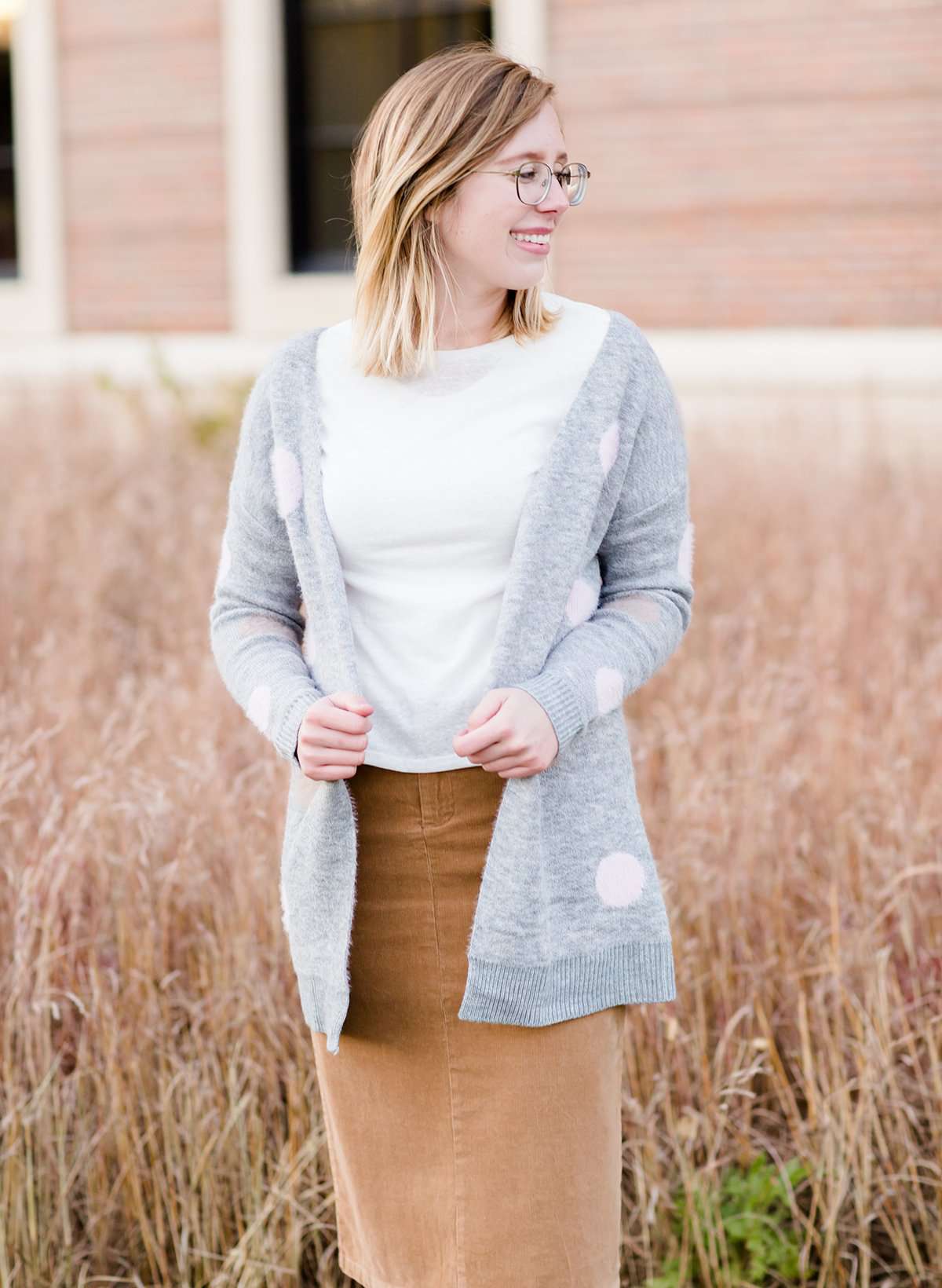 Young woman wearing a modest, long, polka dot, gray, open front cardigan. She is also wearing a khaki corduroy below the knee skirt.