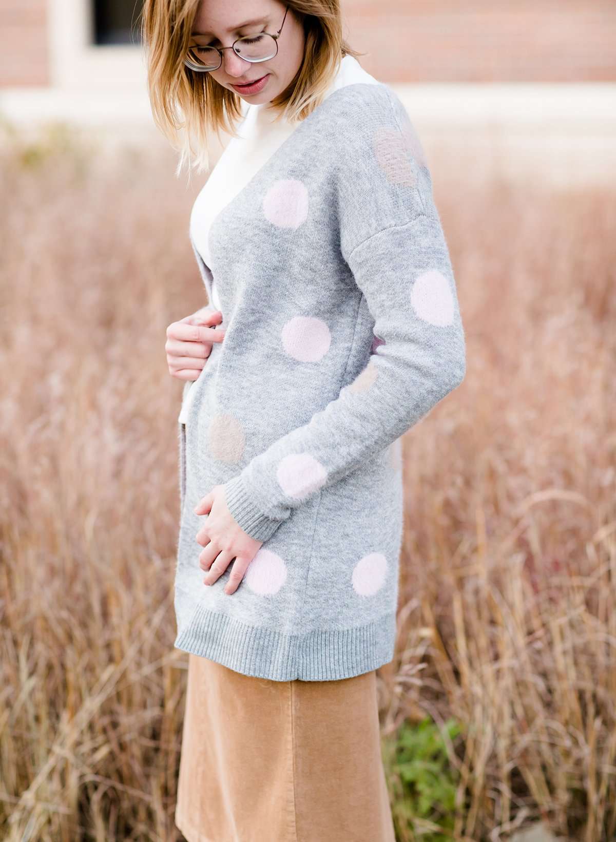 Young woman wearing a modest, long, polka dot, gray, open front cardigan. She is also wearing a khaki corduroy below the knee skirt.