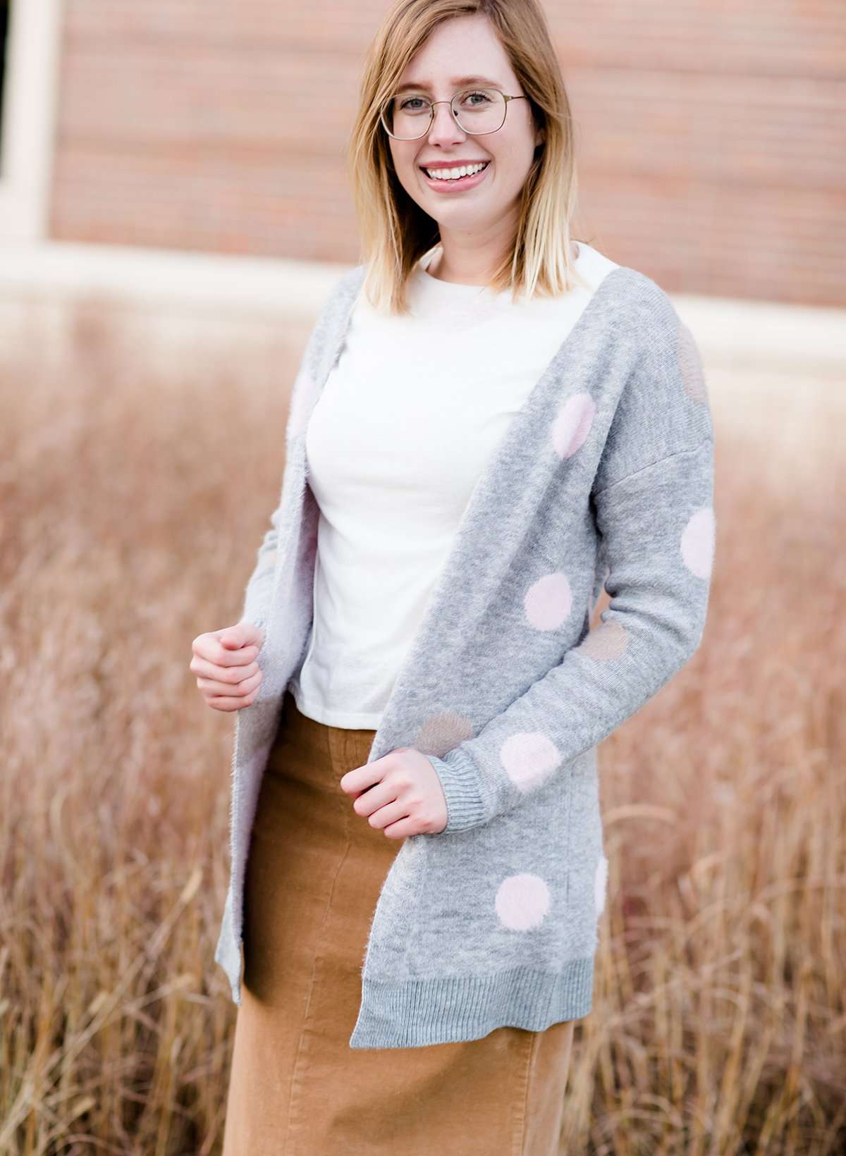 Young woman wearing a modest, long, polka dot, gray, open front cardigan. She is also wearing a khaki corduroy below the knee skirt.