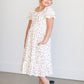 A girl's sized midi length white dress with an allover peach floral design with black stems. It has a high square neckline, puff sleeves, a stretchy waist, and a small ruffle at the hem.