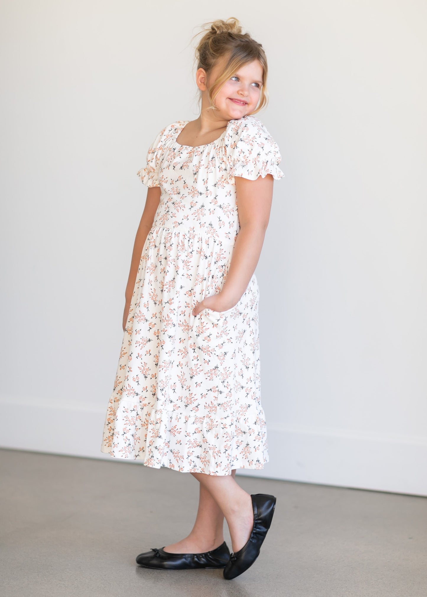 A girl's sized midi length white dress with an allover peach floral design with black stems. It has a high square neckline, puff sleeves, a stretchy waist, and a small ruffle at the hem.
