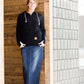 Woman wearing a black, Ampersand half zip, back sweatshirt. It has white toggle strings and a front kangaroo pouch pocket. It is also paired with a long modest jean skirt.