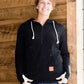 Woman wearing a black, Ampersand half zip, back sweatshirt. It has white toggle strings and a front kangaroo pouch pocket. It is also paired with a long modest jean skirt.