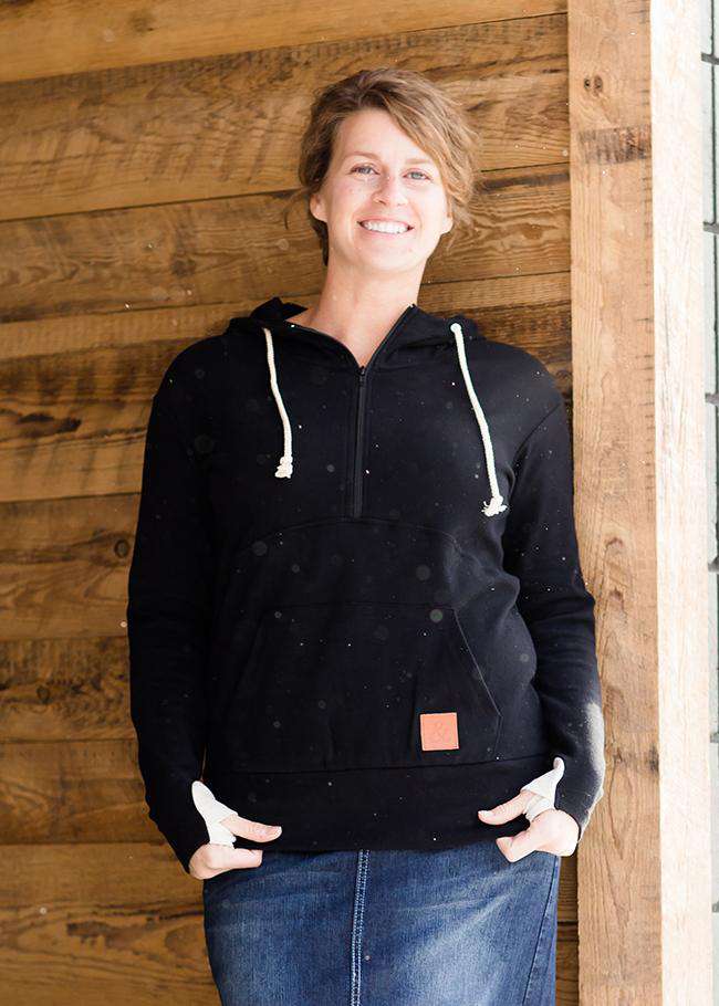 Woman wearing a black, Ampersand half zip, back sweatshirt. It has white toggle strings and a front kangaroo pouch pocket. It is also paired with a long modest jean skirt.