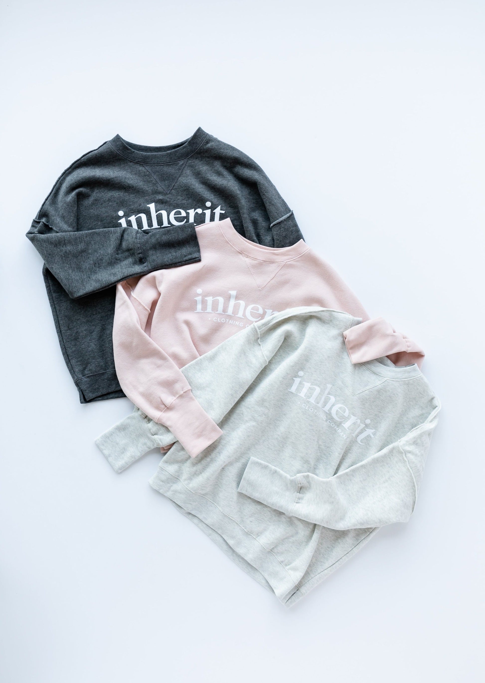Inherit Crew Neck Sweatshirt Tops come in charcoal, blush pink, or hearthered light grey. They have white Inherit logo across the front. There is a raw hem detail on the arms around the bicep and then another raw hem down the shoulder and stopping where the two touch.