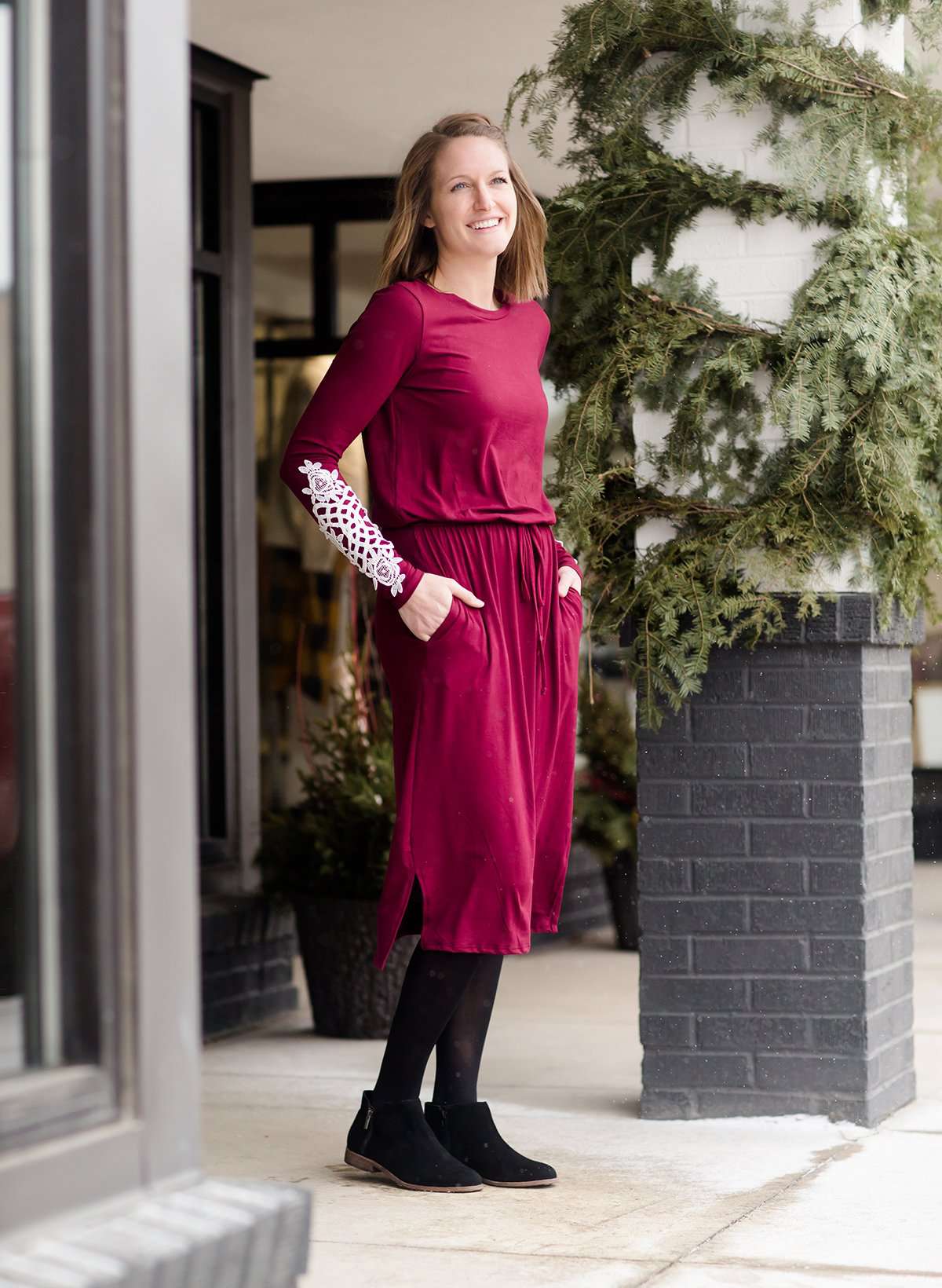 Woman wearing a burgandy and lace midi dress with black tights and sole society back boots. She is standing outside of Inherit Clothing Company.