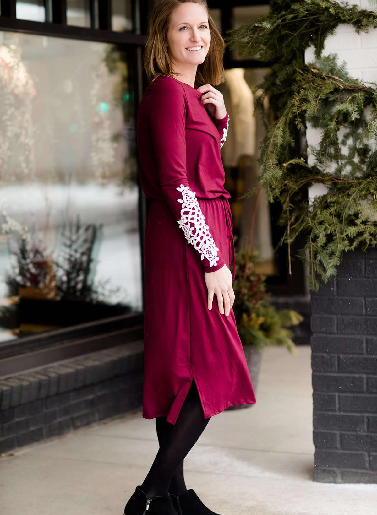 Woman wearing a burgandy and lace midi dress with black tights and sole society back boots. She is standing outside of Inherit Clothing Company.