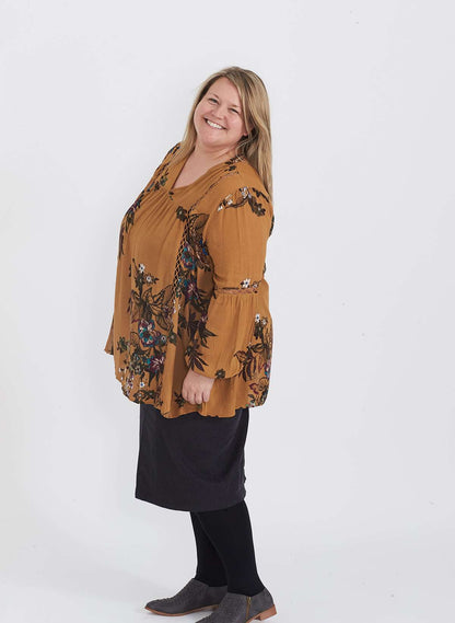 Plus size woman wearing a mustard floral lattice trim style tunic. This top is paired with a below the knee black skirt and leggings.