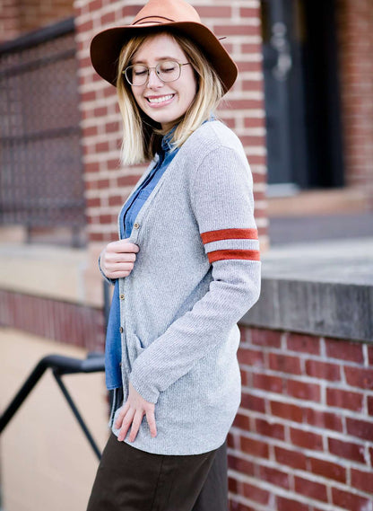 Young woman in hat and skirt wearing a gray open front long knit cardigan with orange stripes on the upper sleeve