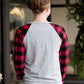 Model woman wearing a grey long sleeve tee with buffalo plaid sleeves and the script font that reads Merry Christmas. 