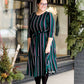 Modest woman wearing a green and black striped midi dress with a self tie bow. She is wearing black leggings and black booties as well. She is at Inherit Clothing Company.