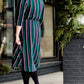 Modest woman wearing a green and black striped midi dress with a self tie bow. She is wearing black leggings and black booties as well. She is at Inherit Clothing Company.