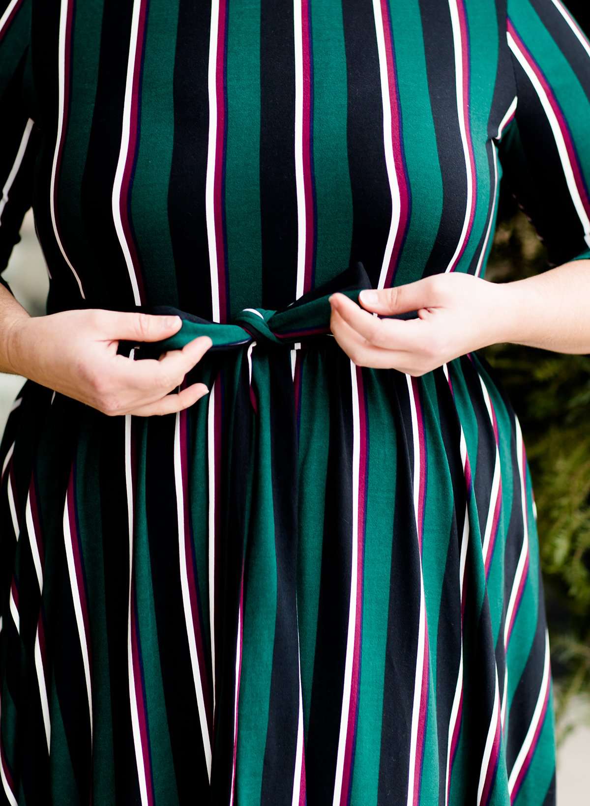 Modest woman wearing a green and black striped midi dress with a self tie bow. She is wearing black leggings and black booties as well. She is at Inherit Clothing Company.