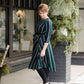 Modest woman wearing a green and black striped midi dress with a self tie bow. She is wearing black leggings and black booties as well. She is at Inherit Clothing Company.
