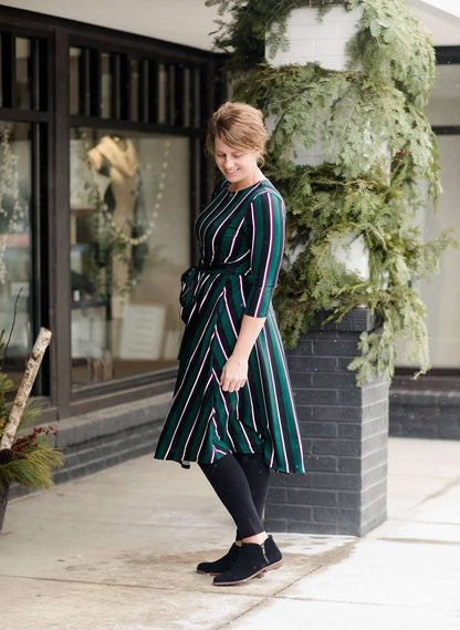 Modest woman wearing a green and black striped midi dress with a self tie bow. She is wearing black leggings and black booties as well. She is at Inherit Clothing Company.