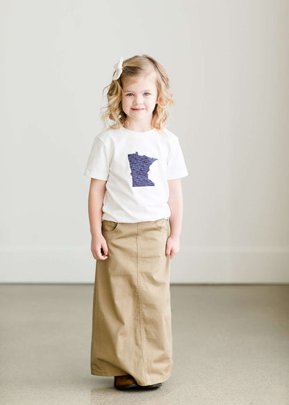 Toddler girl wearing a white minnesota logo on a white tee shirt. She is also wearing a long khaki skirt.