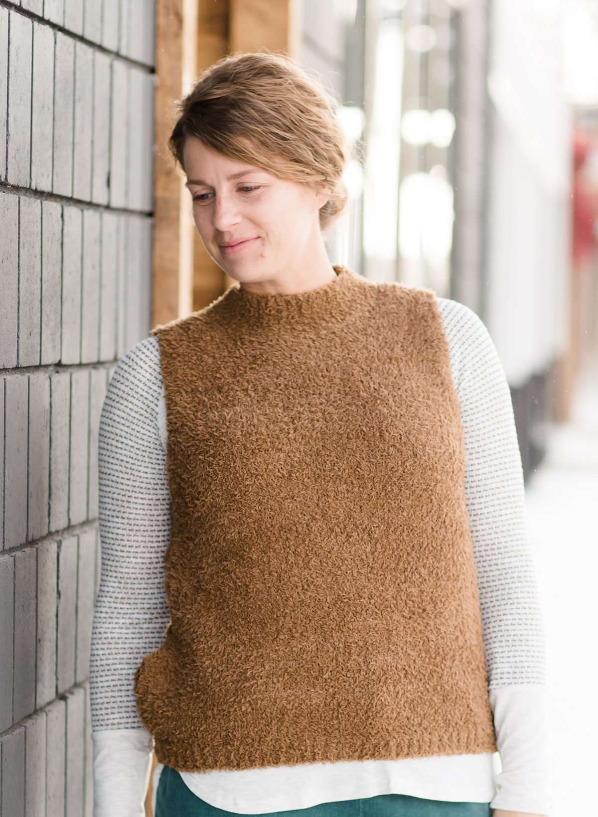 Woman wearing a tan colored fuzzy sweater vest. This vest has open sides and a ribbed bottom. It is also paired with a teal colored below the knee corduroy skirt.