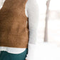 Woman wearing a tan colored fuzzy sweater vest. This vest has open sides and a ribbed bottom. It is also paired with a teal colored below the knee corduroy skirt.