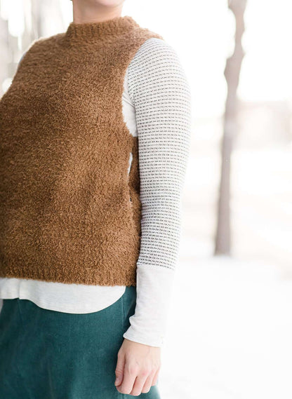 Woman wearing a tan colored fuzzy sweater vest. This vest has open sides and a ribbed bottom. It is also paired with a teal colored below the knee corduroy skirt.