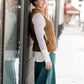 Woman wearing a tan colored fuzzy sweater vest. This vest has open sides and a ribbed bottom. It is also paired with a teal colored below the knee corduroy skirt.