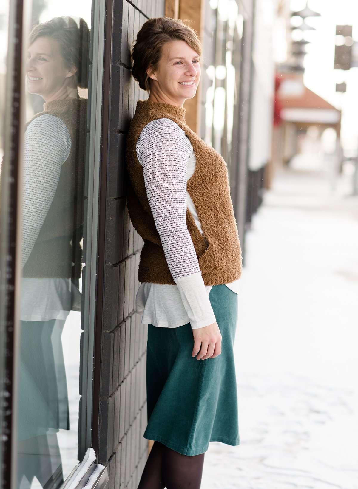 Woman wearing a tan colored fuzzy sweater vest. This vest has open sides and a ribbed bottom. It is also paired with a teal colored below the knee corduroy skirt.