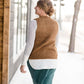 Woman wearing a tan colored fuzzy sweater vest. This vest has open sides and a ribbed bottom. It is also paired with a teal colored below the knee corduroy skirt.