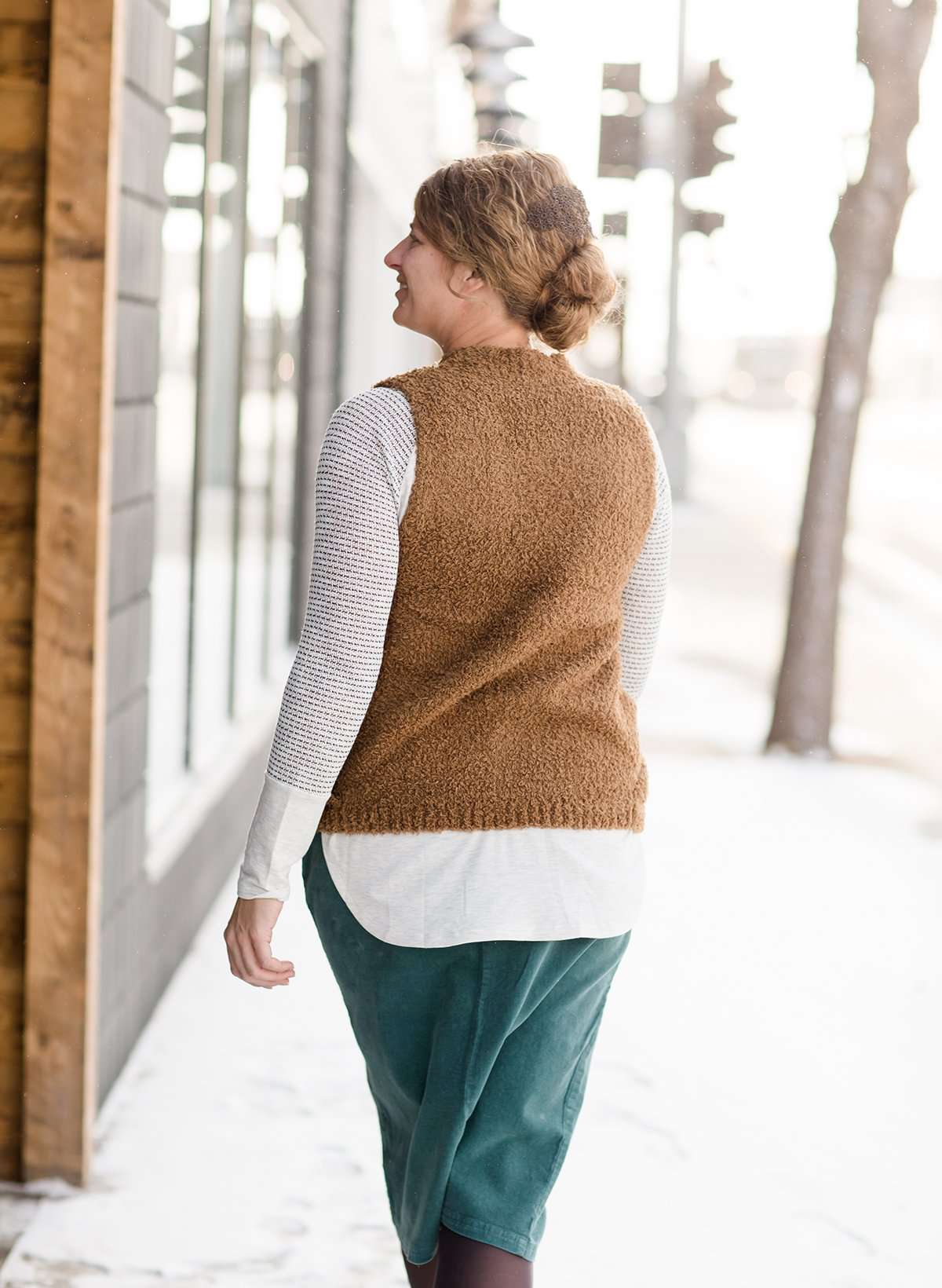 Woman wearing a tan colored fuzzy sweater vest. This vest has open sides and a ribbed bottom. It is also paired with a teal colored below the knee corduroy skirt.