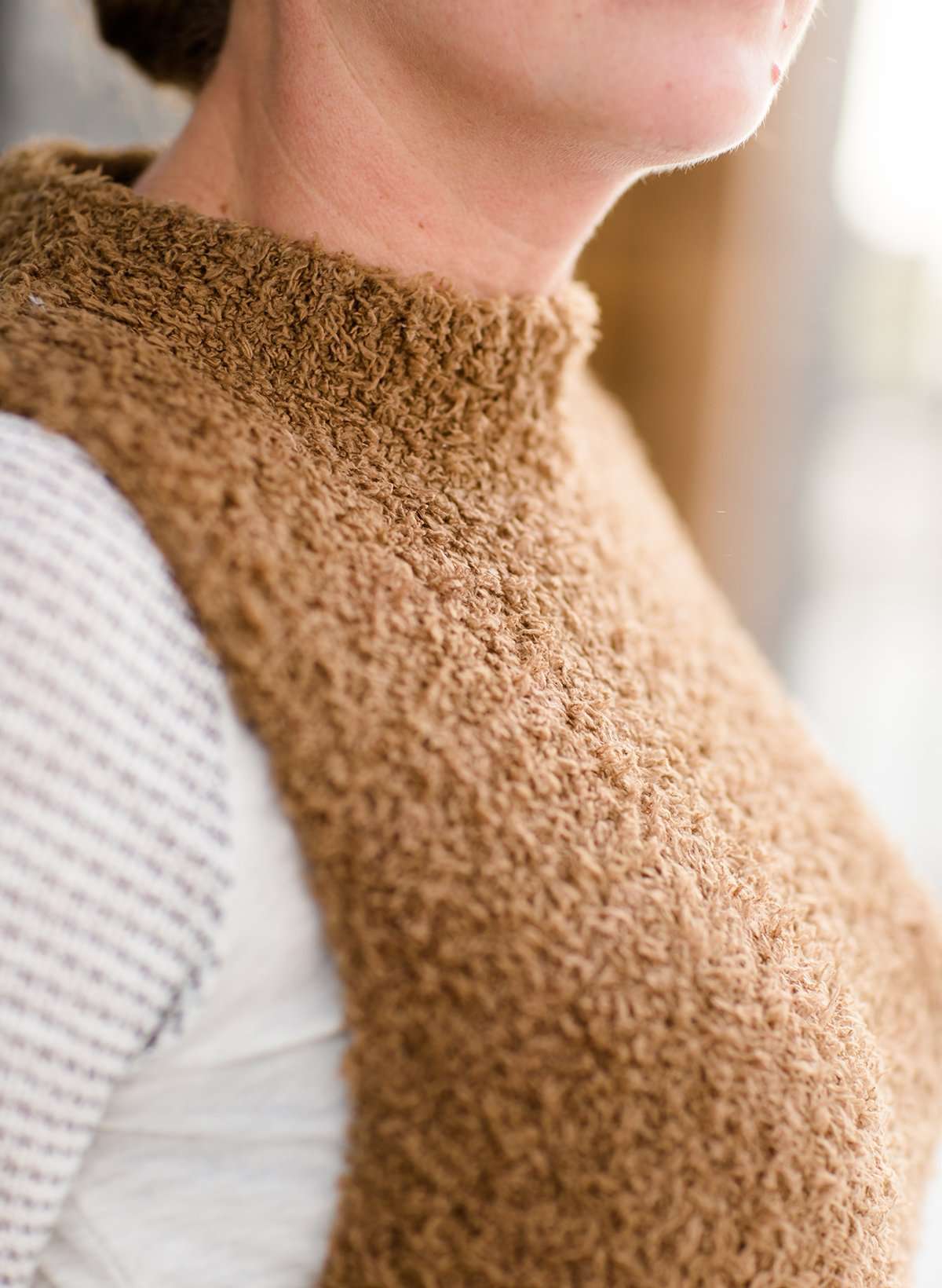 Woman wearing a tan colored fuzzy sweater vest. This vest has open sides and a ribbed bottom. It is also paired with a teal colored below the knee corduroy skirt.