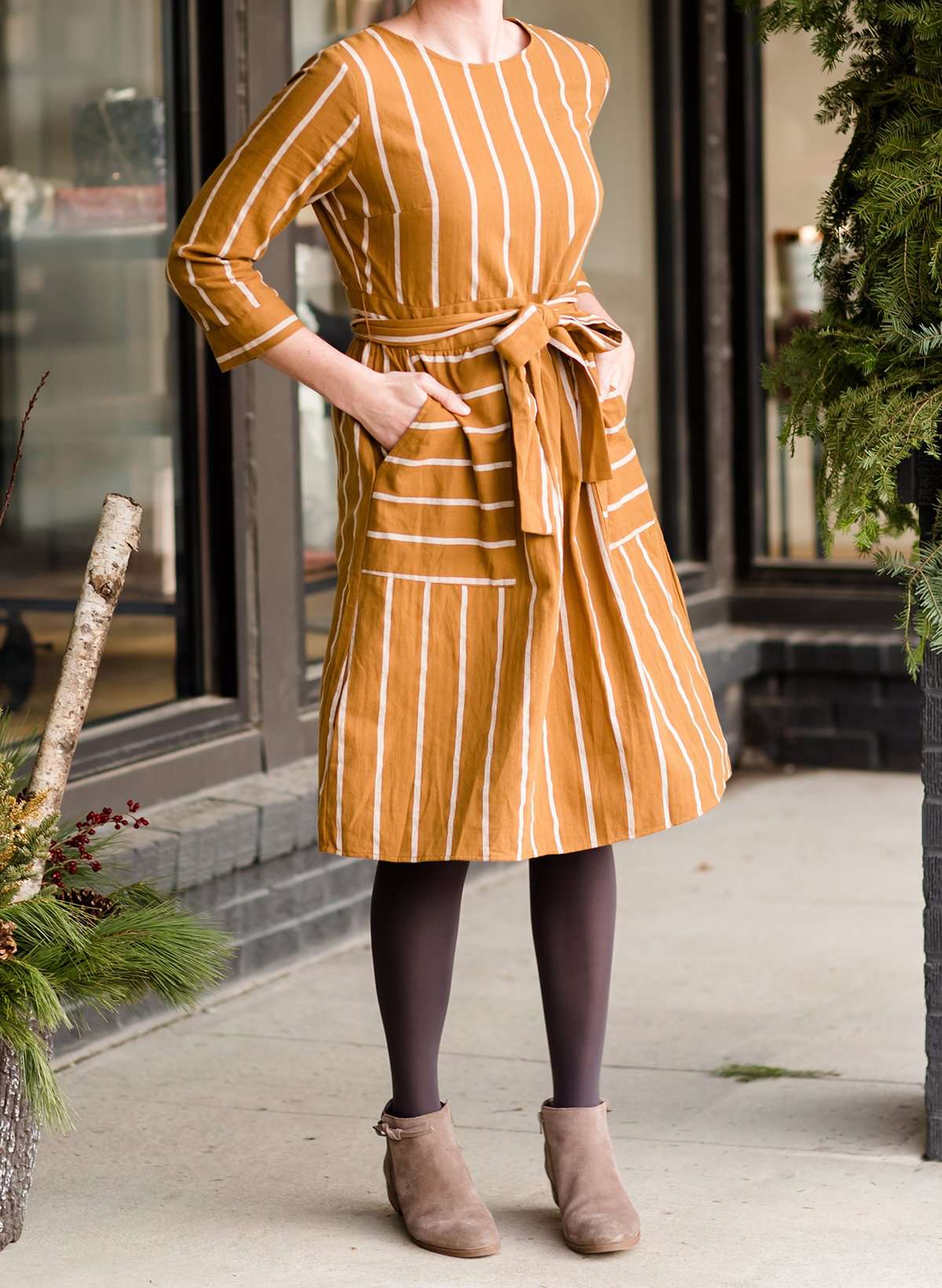 Woman wearing a modest mustard and stripe color midi dress. This dress has oversized pockets a belted front and is paired with tights and brown booties.