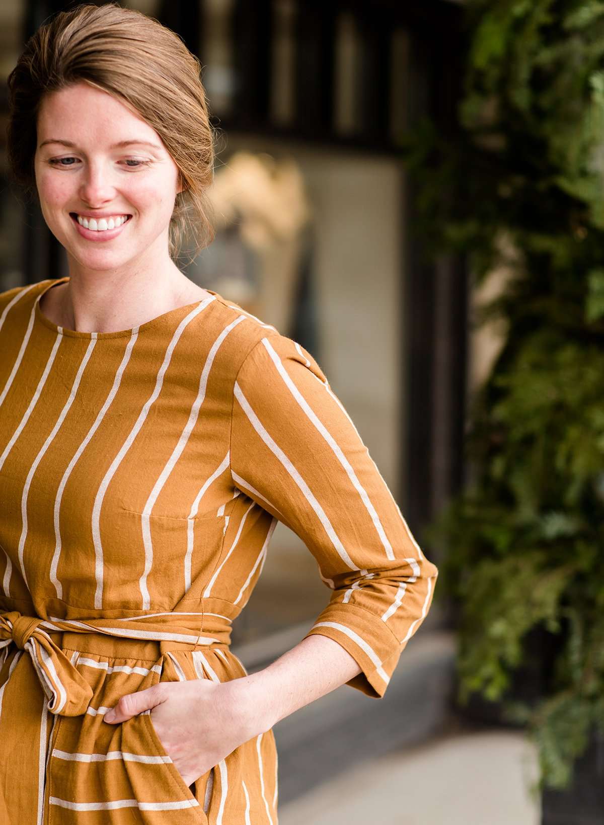 Woman wearing a modest mustard and stripe color midi dress. This dress has oversized pockets a belted front and is paired with tights and brown booties.
