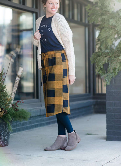 Woman wearing a modest plaid sweater style midi skirt. This skirt has an elastic waist and a tie front. It is paired with tights and short ankle boots.