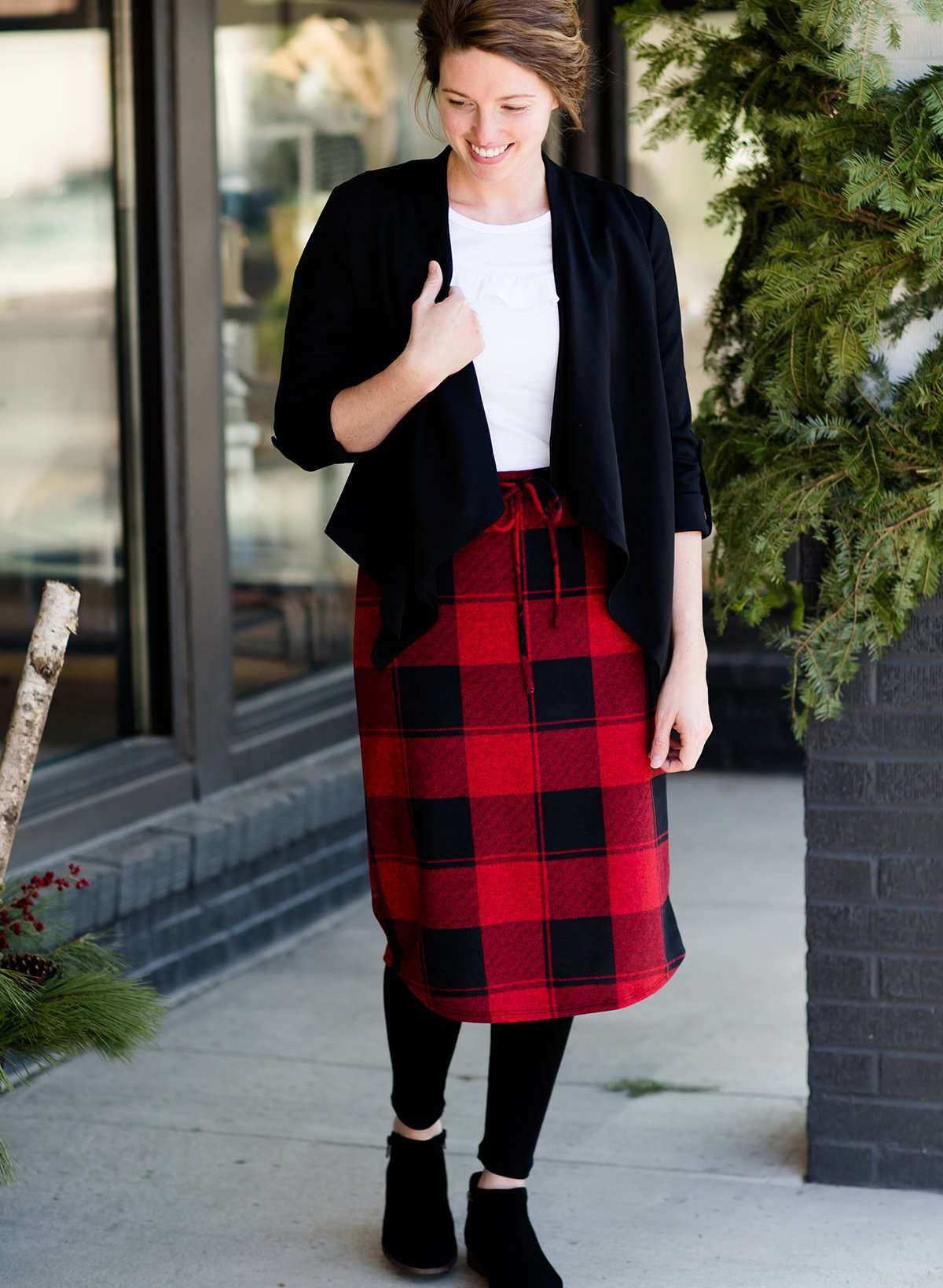  Woman wearing a modest plaid sweater style midi skirt. This skirt has an elastic waist and a tie front. It is paired with tights and short ankle boots.