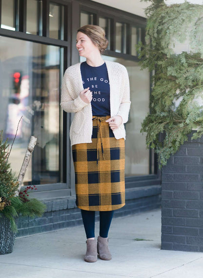 Woman wearing a modest plaid sweater style midi skirt. This skirt has an elastic waist and a tie front. It is paired with tights and short ankle boots.