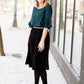 Woman wearing a black, velvet pleated midi skirt. This skirt has a gold accent around the elastic waistband. This modest skirt is paired with a teal, ruffle sleeve poets blouse.