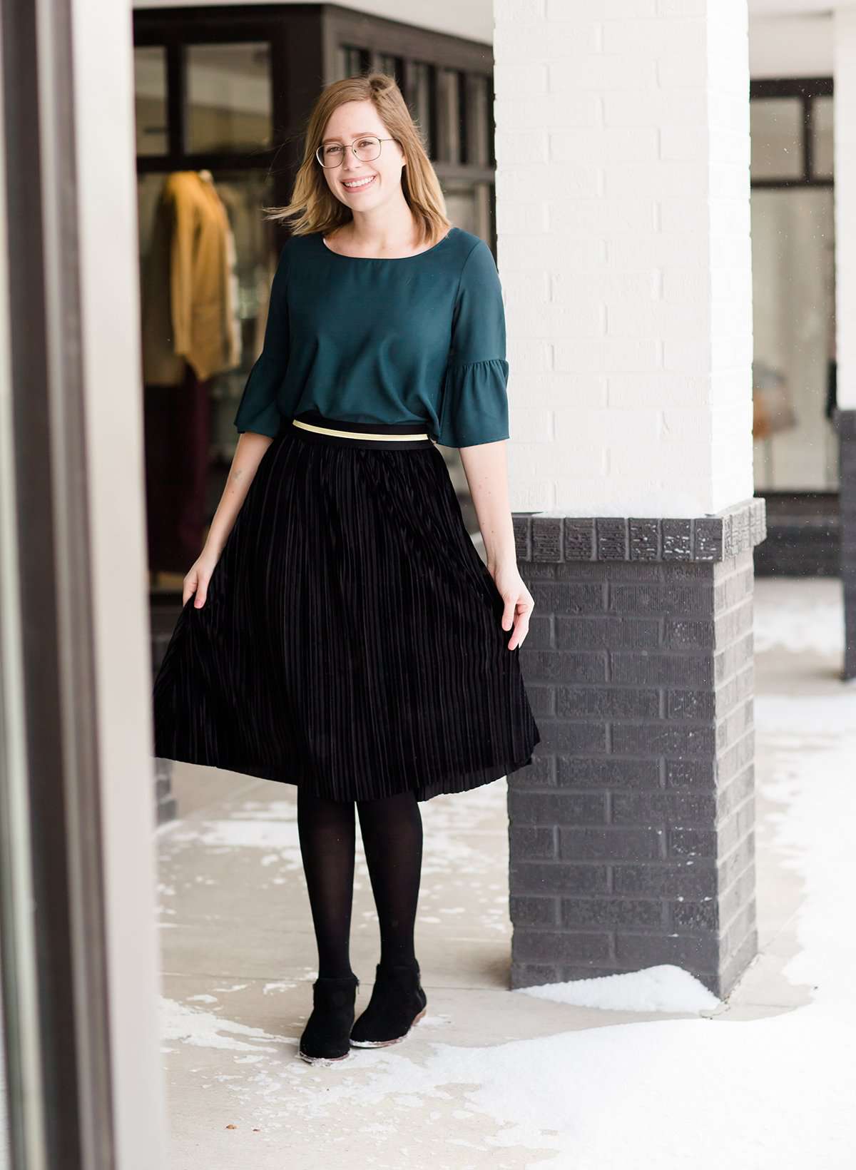 Woman wearing a black, velvet pleated midi skirt. This skirt has a gold accent around the elastic waistband. This modest skirt is paired with a teal, ruffle sleeve poets blouse.