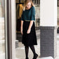 Woman wearing a black, velvet pleated midi skirt. This skirt has a gold accent around the elastic waistband. This modest skirt is paired with a teal, ruffle sleeve poets blouse.