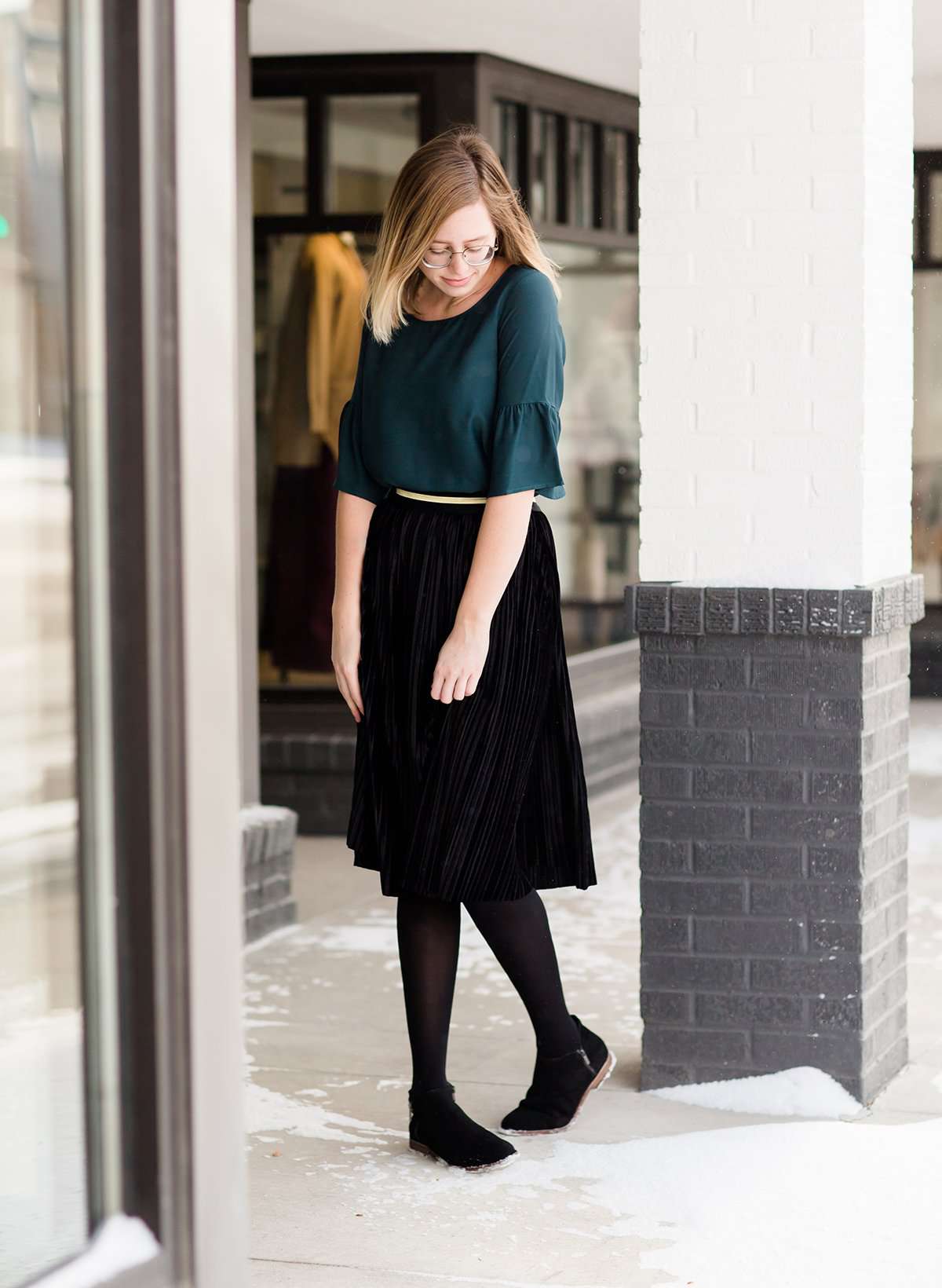Woman wearing a black, velvet pleated midi skirt. This skirt has a gold accent around the elastic waistband. This modest skirt is paired with a teal, ruffle sleeve poets blouse.
