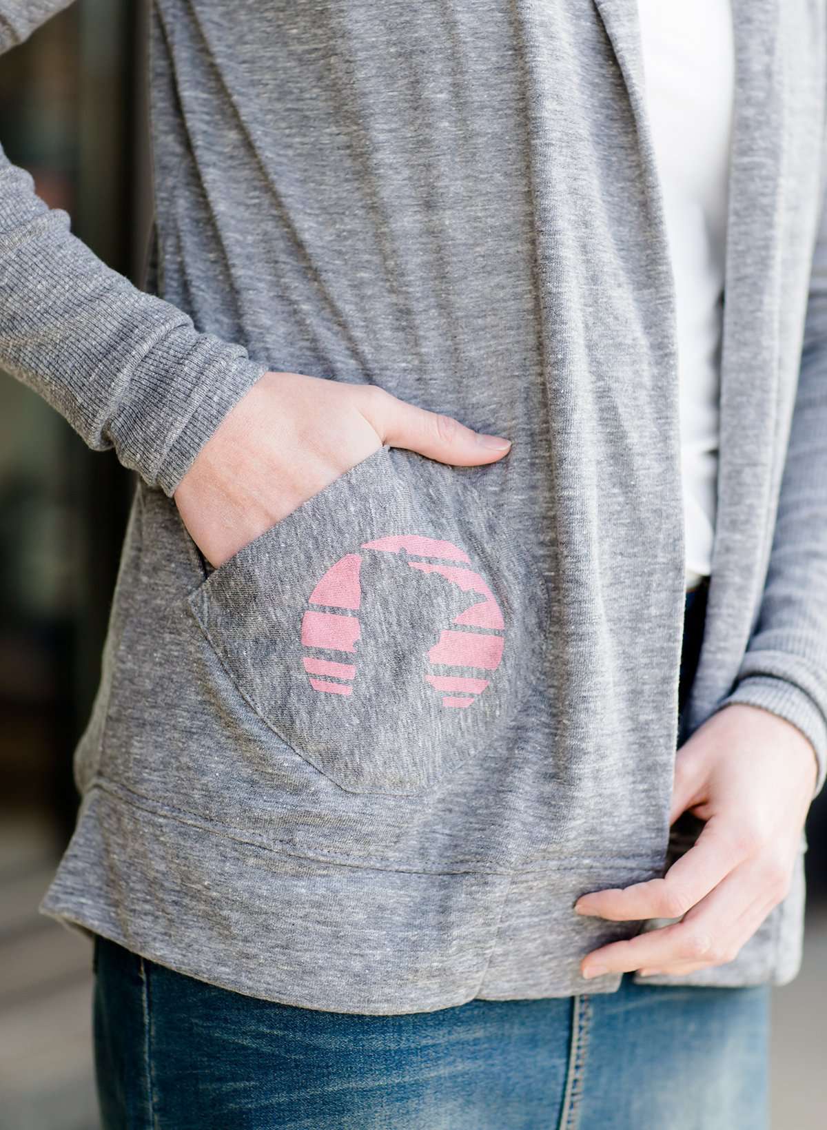 Woman standing in front of Inherit Clothing Company wearing a Minnesota screen printed, gray, open cardigan. The Minnesota screen print is pink.